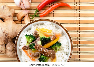 Chinese Cuisine Beef Stir Fry With Vegetables On White Rice, Bamboo Mat Background With Raw Ingredients