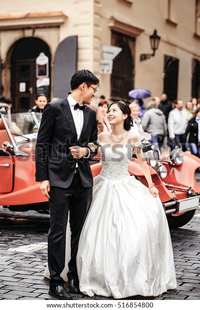 Chinese cute bride and groom\
young newlyweds just married couple on streets of old city on\
wedding day in long white wedding dress stands outdoors near red\
retro car