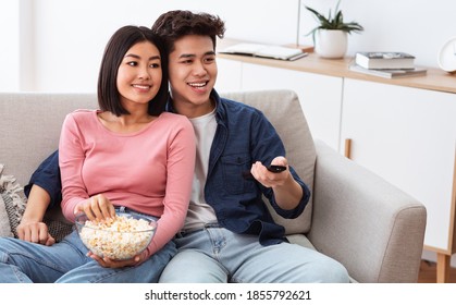 Chinese Family Couple Watching TV Together And Eating Popcorn On Weekend, Switching Television Channels With Remote Control Sitting On Couch In Living Room At Home.