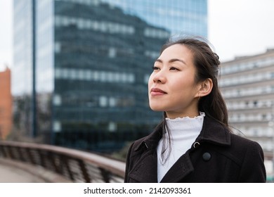 Chinese Female Entrepreneur In Black Coat Looking Away And Observing City Street While Standing On Bridge Against Skyscraper In Daytime In Downtown