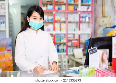 Chinese Female Pharmacist In Protective Facial Mask Standing Among Shelves In Drug Store. High Quality Photo