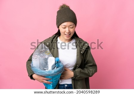 Chinese girl holding a bag full of plastic bottles to recycle over isolated pink background smiling a lot