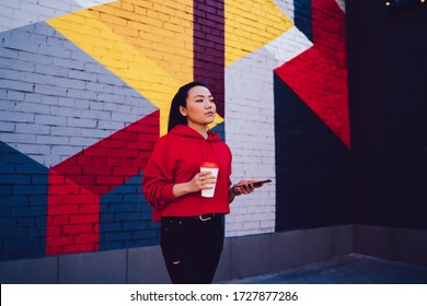 Chinese influence blogger holding modern cellphone device and coffee to go in hands and thinking on publication idea, pensive millennial with smartphone pondering near colorful wall with street art