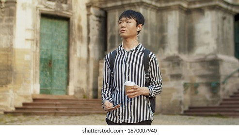 Chinese Male Student Tourist In Headphones Listening To Music On Smartphone And Drinking Coffee Young Asian Man Having Sightseeing Tour Around City And Sipping Drink. Guy Walking Town While Travelling