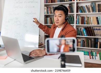 Chinese Male Tutor Having Online Lecture Via Video Call On Phone Pointing At Whiteboard Sitting At Desk In Library Indoors. E-Teaching Concept. Selective Focus On Teacher