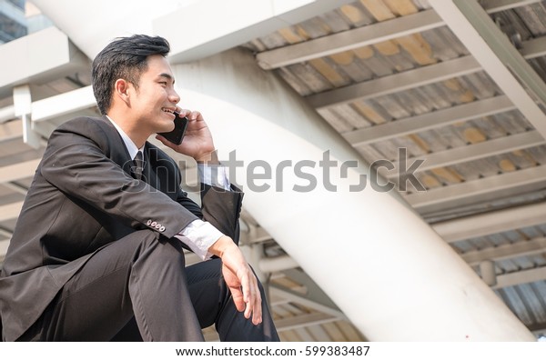 Chinese man wears\
business suit is talking on mobile phone. He is at the station. He\
is sitting at stairs.