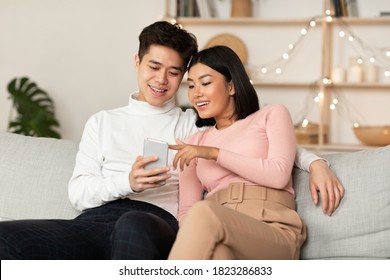 Chinese Millennial Couple Using Smartphone Relaxing On Weekend Sitting On Couch At Home, Spending Evening Together. Mobile Application, Phone Users Browsing Internet, Gadget Lifestyle Concept