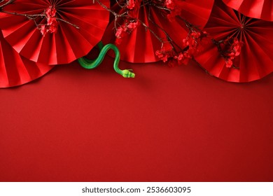 Chinese New Year background with decorative paper fans, cherry blossoms, and a green snake toy on a red background. Ideal for festive and cultural themes - Powered by Shutterstock