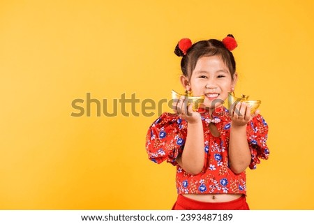Chinese New Year. Happy Asian Chinese little girl smile wearing red cheongsam holding gold ingot, Portrait children in traditional dress hold golden bar, studio short isolated on yellow background
