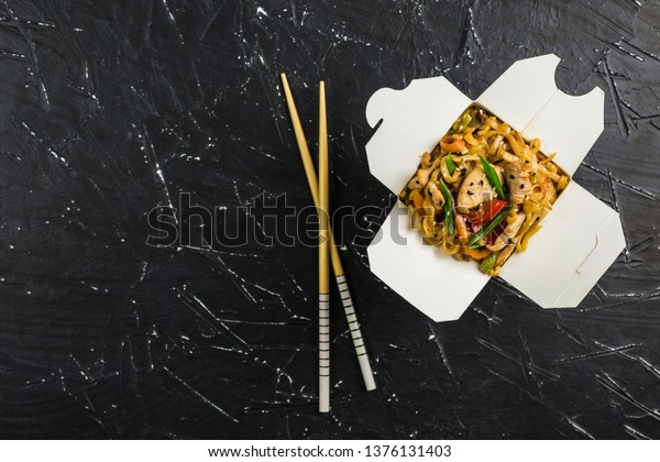 Chinese noodles in a box with chicken and\
vegetables with sticks. Wok food delivery from restaurant on dark\
background from top\
view.