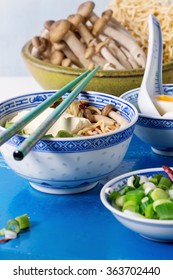 Chinese porcelain bowl of asian ramen soup with feta cheese, noodles, spring onion and mushrooms, served with turquoise chopsticks and nboiled egg over white kitchen table with blue stone board. 