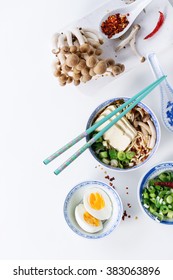 Chinese Porcelain Bowl Of Asian Ramen Soup With Feta Cheese, Noodles, Spring Onion And Mushrooms, Served With Turquoise Chopsticks And Sliced Egg Over White Background. Top View