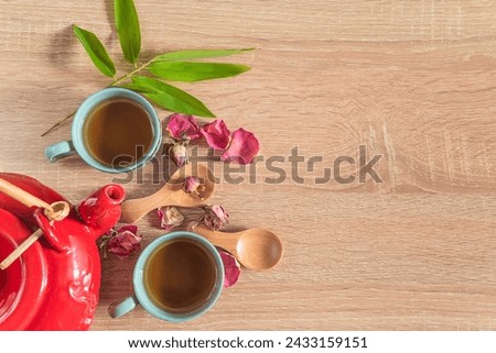 Chinese tea time concept: two chineese cups of tea, red teapot, dry petals of red rose flowers and bamboo leaves on a wooden background; copy space