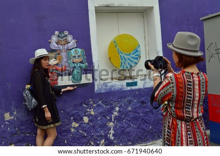 Chinese traveler girl make a photo or video on her camera of the famous place in Penang, Malaysia - Graffiti, street art on the wall and her friend. Stylish tourists have a nice dresses and hats.