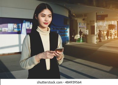 Chinese Woman CEO Is Waiting For An Answer On Her Text Message On Mobile Phone, While Is Standing In Airport Terminal Before Her Business Trip Abroad. Hipster Girl Is Using Cell Telephone For Chat