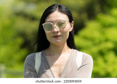 A Chinese Woman With Closed Lips Standing Against A Blurred Green Background Outside On A Sunny Day.