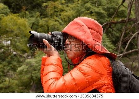 Chinese woman dressed in winter clothing taking photographs with a Nikon D3100 camera, holding camera correctly and looking through the viewfinder.