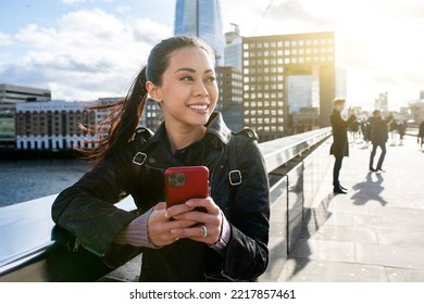 Chinese Woman In London Smiling And Using Mobile Phone In The City - Young Asian Woman On London Bridge In The City Holding A Smartphone And Looking Away - Communication And Lifestyle Concepts