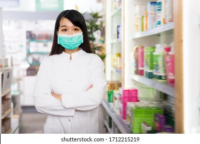 Chinese Woman Pharmacist In Protective Facial Mask Standing With Arms Crossed In Interior Of Pharmacy