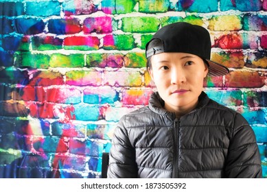 A Chinese Woman Posing Against A Color Brick Background Wearing A Jacket And Baseball Hat Worn To The Side While Looking Away.