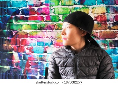 A Chinese Woman Posing Against A Color Brick Background Wearing A Jacket And Baseball Hat Worn To The Side While Looking Away.