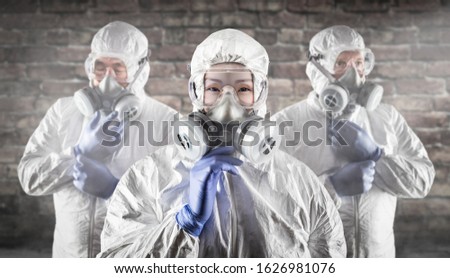 Chinese Woman and Team Behind In Hazmat Suites, Gas Masks and Goggles Against Brick Wall.