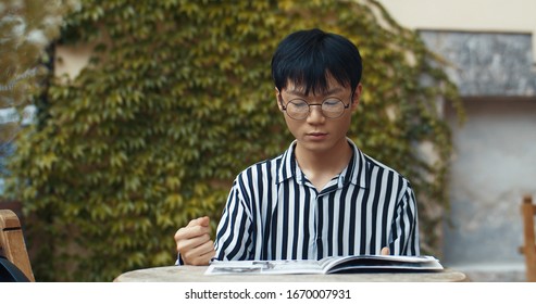 Chinese Young Man In Glasses Reading Magazine And Flipping Pages While Sitting At Table In Cafe Terrace Outdoors. Asian Guy Student Looking Through Media Outside.