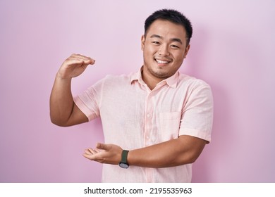 Chinese young man standing over pink background gesturing with hands showing big and large size sign, measure symbol. smiling looking at the camera. measuring concept. 
