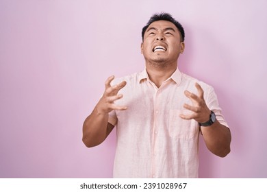 Chinese young man standing over pink background crazy and mad shouting and yelling with aggressive expression and arms raised. frustration concept. 
