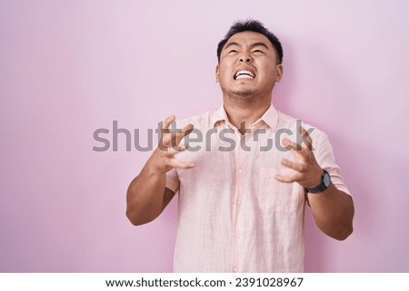 Chinese young man standing over pink background crazy and mad shouting and yelling with aggressive expression and arms raised. frustration concept. 