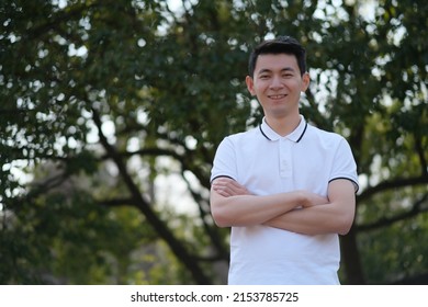 Chinese Young Man In White Short Sleeve Polo Shirt, Cross His Arms, Smile And Looking At Camera In Green Park