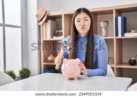 Chinese young woman holding hammer and piggy bank puffing cheeks with funny face. mouth inflated with air, catching air. 
