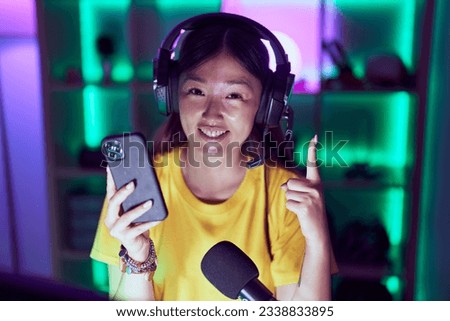 Chinese young woman playing video games with smartphone smiling with an idea or question pointing finger with happy face, number one 