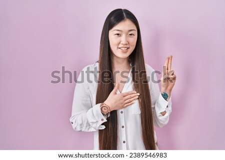 Chinese young woman standing over pink background smiling swearing with hand on chest and fingers up, making a loyalty promise oath 