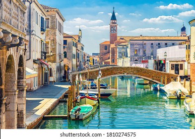 Chioggia cityscape with narrow water canal with moored multicolored boats, old buildings, brick bridge and tower of San Giacomo Apostolo church, blue sky in summer day, Veneto Region, Northern Italy
