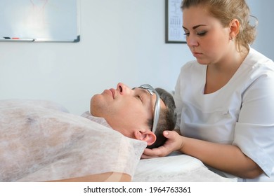 Chiropractor Doctor Is Putting On Electronic Headband From Migraine On Man's Forehead. Treatment Of Headaches With Electric Current In Clinic On Procedure With Manual Physiotherapist.