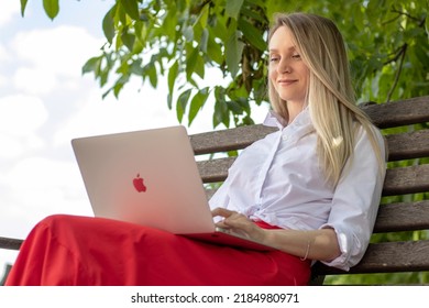 Chisinau,Moldova - 07.31.2022:young pretty young woman working on laptop macbook air sitting on park bench under a walnut tree.blonde smiling business lady is smiling and typing on keyboard