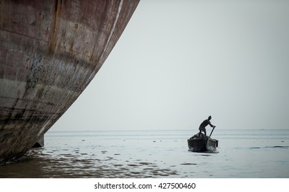 Chittagong, Bangladesh - circa February 2016:  man rowing a small boat in between big ocean ships that are being broken down in a ship breaking yard in Chittagong