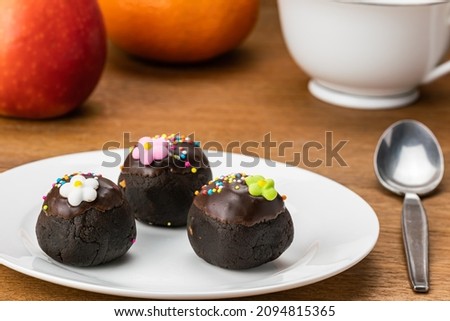 Choc balls or chocolate balls topping with multicolored rainbow sprinkles and colored sugar flower in white ceramic dish on wooden table with red apple, ripe orange and a cup of coffee.