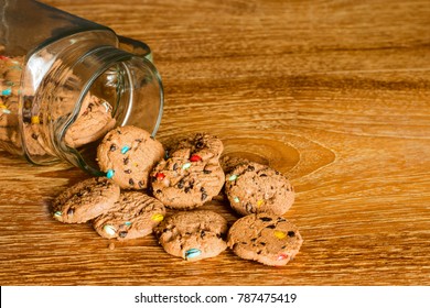 Choc Chip Smartie Cookies Falling Out Of Jar On Wooden Table Top.