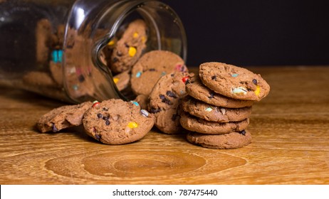 Choc Chip Smartie Cookies Falling Out Of Jar On Wooden Table Top.