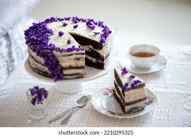 Choclade Cake Layered With Vanilla Cream, Decorated With Flowers Violets. A Piece Of Cake Cut Out, Lying On A Plate , Two Dessert Forks Next To It, And A Cup Of Tea On The Right. Few Flowers Sprinkled