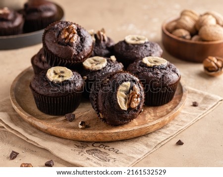 Chocolate banana muffins with chocolate pieces and chopped walnuts on wooden plate. Beige background, textile napkin. Close up food. Homemade brownie dessert. 