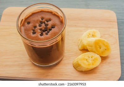 Chocolate Banana Shake Topping Sprinkled With Chocolate Chips In A Glass, Next To A Sliced Banana All On A Wooden Cutting Board.