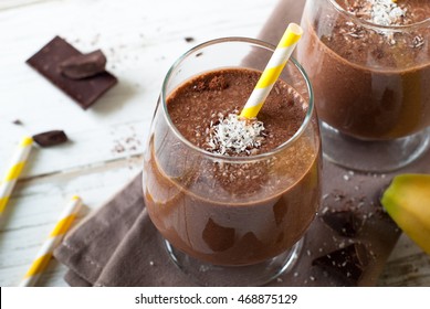 Chocolate banana smoothie or milkshake with coconut on white wooden table.