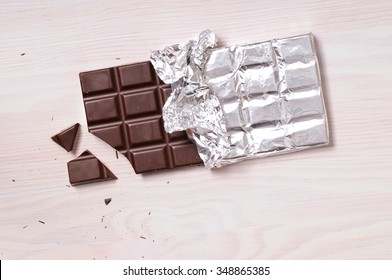 Chocolate Bar With Silver Wrapping On A Wooden Table With A Broken Portion. Horizontal Composition. Top View