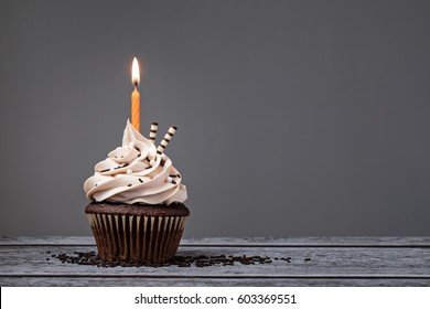 Chocolate Birthday Cupcake With  Buttercream Icing And Orange Candle Over A Grey Background.