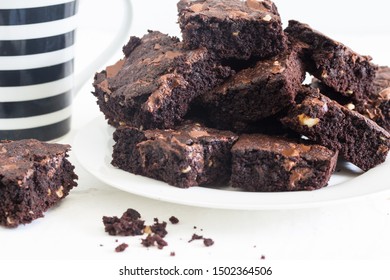 Chocolate brownies in a stack on white plate with black and white defocused coffee mug in background on white background