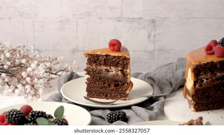 Chocolate cake slice with caramel frosting and a raspberry on top, surrounded by fresh berries and a floral arrangement, creating a cozy dessert scene - Powered by Shutterstock