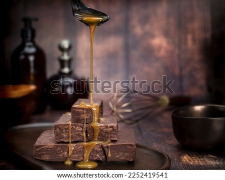 Chocolate caramel fudge stacked on a plate with caramel sauce pouring over the top.  Sauce being poured from a spoon. Dark kitchen objects in the background with vintage wood boards.